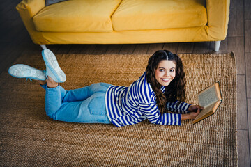 Lovely woman lying on rug with a book in cozy home interior next to a sofa, enjoying leisure time,...