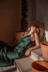 young blonde teen girl in green silk dress is sitting casual like cowboy with straw hat on head and looking up on the italian patio background at sunny day. travel lifestyle concept, free space