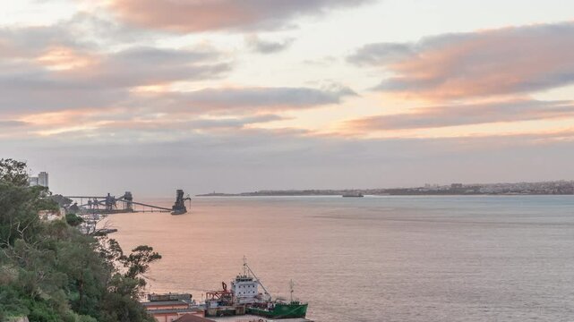 Aerial timelapse from a hill near Porto Brandao Ferry Station on the Tagus River in Lisbon. Dramatic sunset with colorful clouds, a ship floating on water and Terminal Portuario Trafaria in background