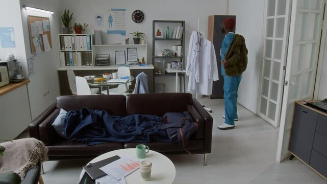 Young male clinician in medical scrubs and jacket entering break room, putting backpack on leather couch, sitting down and taking out plastic container with food and bottle with hot tea