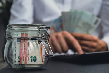 Man hands counting expenses banknotes of euro cash from glass jar in the piggy bank on calculator....