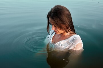 A serene portrait of a young woman in a wet white shirt standing waist-deep in calm water, wearing a cross necklace and creating ripples, conveying peace and contemplation in nature