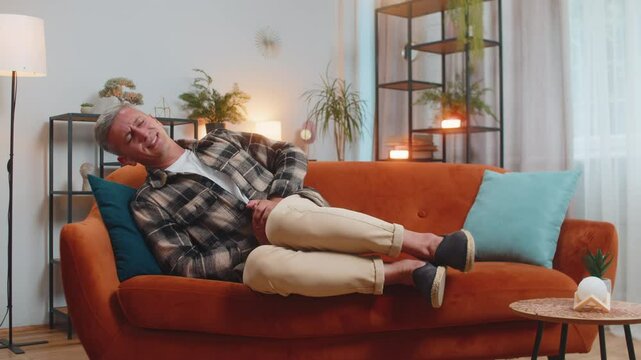 Young Caucasian man lying on sofa feeling sudden strong abdominal stomach ache, gastritis problem. Guy having symptom poisoning diarrhea indigestion peptic ulcer pancreatitis at home apartment room.