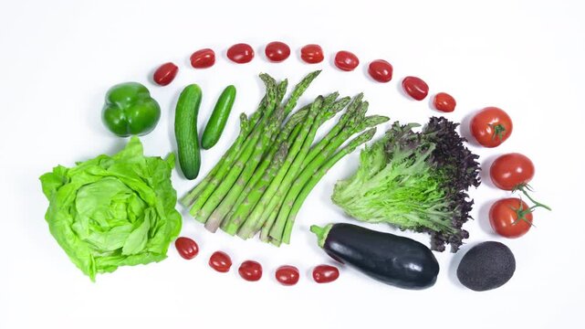 Green and purple vegetables surrounded by red cherry tomatoes on white background. Asparagus, lettuce, cucumbers, peppers, eggplant and avocado. Proper nutrition. Vegetarianism and veganism concept.