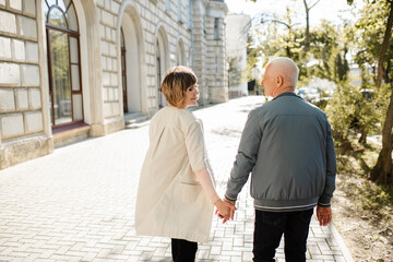 Senior happy couple of stylish man and woman 60-65 year old walking together holding hands on streets at city outdoors rear view. Wellness lifestyle.