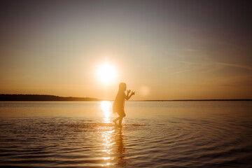 Silhouette of a girl running on water against the background of an evening sunset. Water splashes in all directions. Carefree baby fun. Happy healthy childhood.