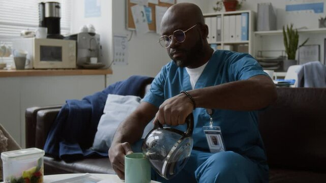 Young tired male doctor in eyeglasses and medical scrubs sitting on couch in break room after work, pouring hot tea in mug, drinking it and leaning on back of sofa