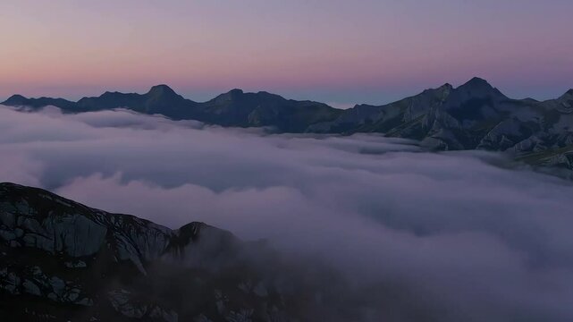 Ocean of clouds rolling over rugged mountains under a purple dawn sky.