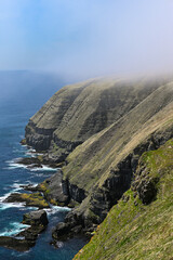 A grassy green cliff's edge of a high mountain with the ocean in the distance. A thick mist of fog crept up over the lush land. The ocean's coastline is rocky with waves crashing onshore. 