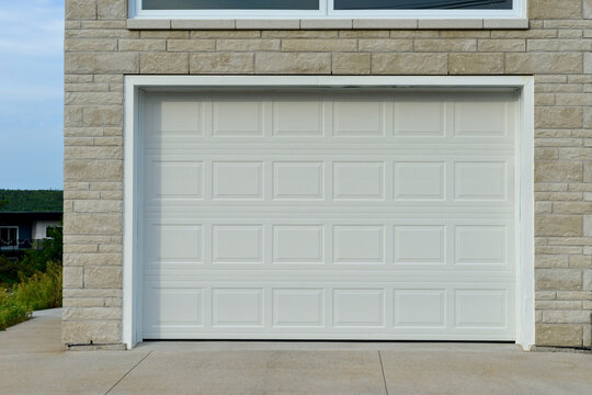 A large white metal car garage door on a grey brick house. The solid single steel door has square patterns or panels. The modern luxury automotive building has a grey concrete driveway entrance. 