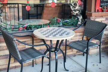 A black metal bistro table with two black resin wicker chairs. The empty outdoor dining set is on a concrete sidewalk next to a beige brick wall and patio railing. The coffee shop dining area is empty