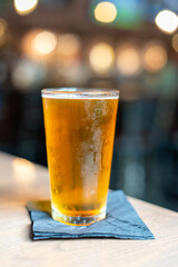 A pint glass sits on a napkin filled with a pale ale craft beer. The liquid has a yellow tint. A clear beer glass sits on a table at a microbrewery. The background has multiple bokeh blurry lights.