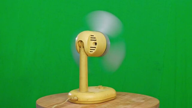 A vintage-style electric fan with blurred, rotating blades sits on a wooden surface against a bright green screen background. The fan's blades are in motion, creating a sense of airflow