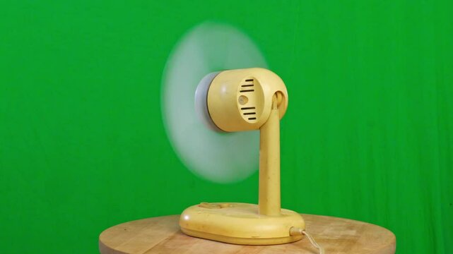 A vintage-style electric fan with blurred, rotating blades sits on a wooden surface against a bright green screen background. The fan's blades are in motion, creating a sense of airflow
