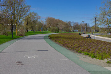 A scenic park pathway in Tallinn curves through lush greenery under a clear blue sky. The bike lane, marked with a bicycle symbol, is flanked by green grass, trees, and benches. Modern street lamps