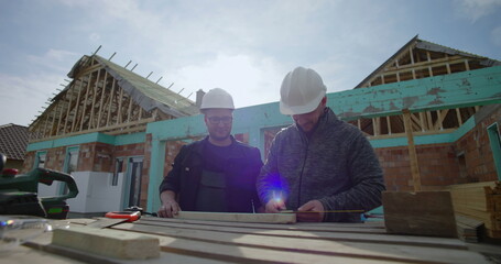Fototapeta premium Builders marking wood on a construction site, teamwork and focus on details, residential project, hard hats, tools and materials on the worktable, house under construction