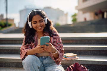 Fototapeta na wymiar Happy Indian student using her cell phone while listening music over headphones at campus.