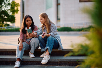 Fototapeta premium Happy female students using cell phone while relaxing on staircase at campus.