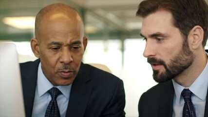 Two men, one Black and one White, engage in a discussion about work strategies in a contemporary office environment. Their focused expressions indicate a serious conversation about projects