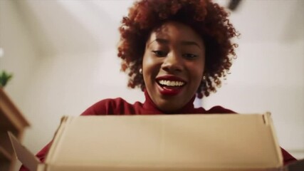 A young Black woman expresses pure joy while opening a surprise package in her cozy living room. Her excitement catches the viewer's attention in this delightful moment