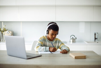 Seven year old mixed race boy doing homework with laptop and headphones at modern kitchen.