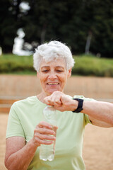 An elderly woman opens a bottle of water after exercising in a park.