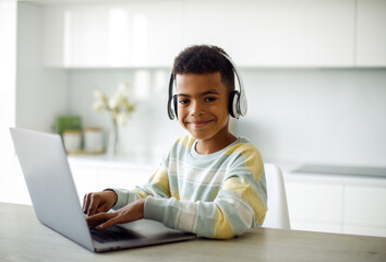 Seven year old mixed race boy doing homework with laptop and headphones at modern kitchen.
