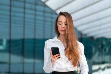Elegant businesswoman with long brown hair using smartphone outside modern office building. Beautiful woman is in white blouse conveying professionalism and focus