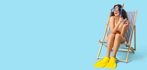 Young African-American woman in snorkeling mask and flippers sitting on deck chair against blue background with space for text © Pixel-Shot