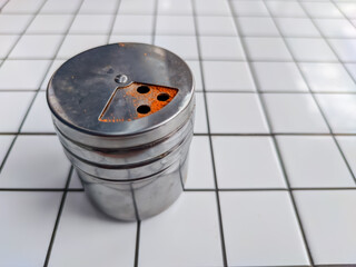 A silver chili powder dispenser sits on a black-and-white checkered tile surface. Reddish residue near the dispensing holes indicates its contents.