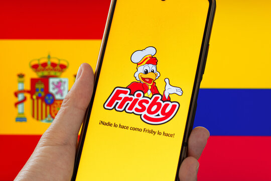 Hand holding a cell phone with the Frisby app open in front of the flags of Spain and Colombia, alluding to the international dispute over the brand. Restaurant chain of Colombian origin