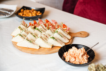 Party Sandwich Platter with Meat and Spread. Assorted triangle sandwiches with cold cuts and greens served on a wooden board, alongside a creamy spread and snacks.