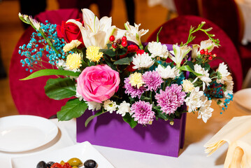 Colorful Flower Arrangement in Purple Box Vase. Bright mixed flower bouquet with roses, lilies, and chrysanthemums in a purple container on a banquet table setting.