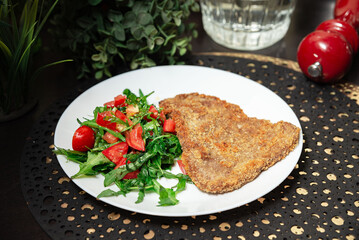 Breaded schnitzel with fresh tomato arugula salad. Crispy breaded schnitzel served with a side of fresh arugula and chopped tomato salad on a white plate.