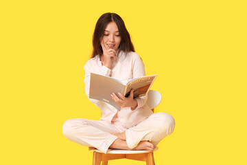 Beautiful young Asian woman in pajamas with magazine sitting on chair against yellow background