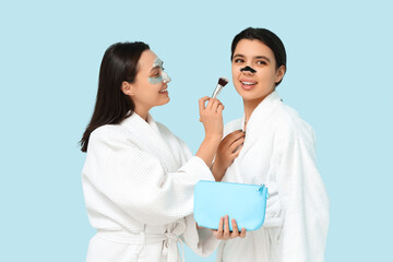 Beautiful young women in bathrobes making cosmetic procedures to each other on color background