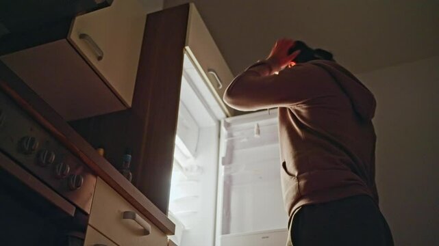 A man opens an empty refrigerator at night and looks into it, finds nothing and closes the refrigerator. An individual is reaching into an open refrigerator located in a modern, stylish kitchen