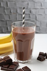 Tasty chocolate milk in glass, pieces and bananas on white wooden table, closeup