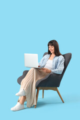 Beautiful woman using laptop in armchair on blue background