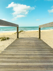 Wooden boardwalk leads to a sunny beach with gentle waves and a clear blue sky dotted with fluffy...