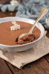 A creamy chocolate mousse in a white bowl, with a spoonful lifted, is delicately garnished with white flowers and rests on parchment paper atop a wooden surface