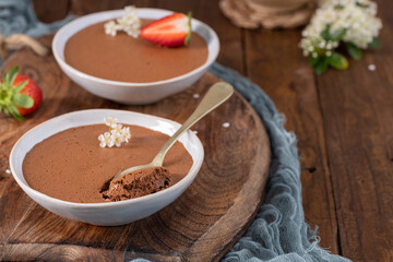 Individual bowls of rich chocolate mousse, one with a spoonful lifted, garnished with strawberries and delicate white flowers, arranged on a wooden board with soft blue fabric