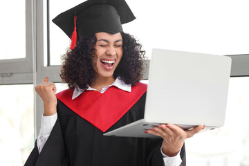 Happy African-American female graduate with laptop showing yes gesture near window
