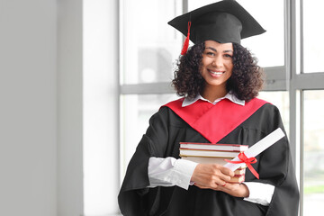 Happy African-American female graduate with diploma and books near window