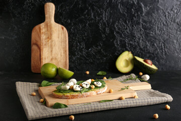 Tasty sandwich with avocado hummus, mozzarella cheese, basil, wooden board and napkin on dark...