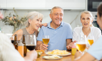 Group of joyful elderly women and man gathered around cozy kitchen table, chatting amiably while...