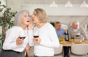 Two elderly women friends chatting and drinking wine in kitchen at home