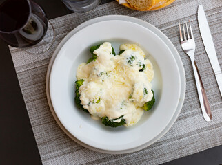 Appetizing broccoli and cauliflower gratin garnished with cheese and cream served on white plate near glass of wine and bread.