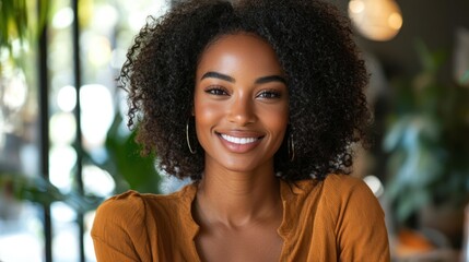 A young woman with curly hair smiling at the camera in a cozy, green-painted room with a wooden table and potted plants.