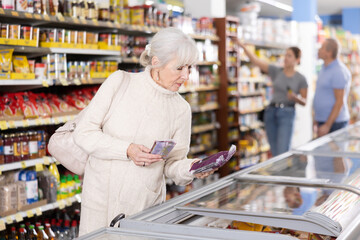Watchful aged woman customer selecting frozen food from market fridge in superstore